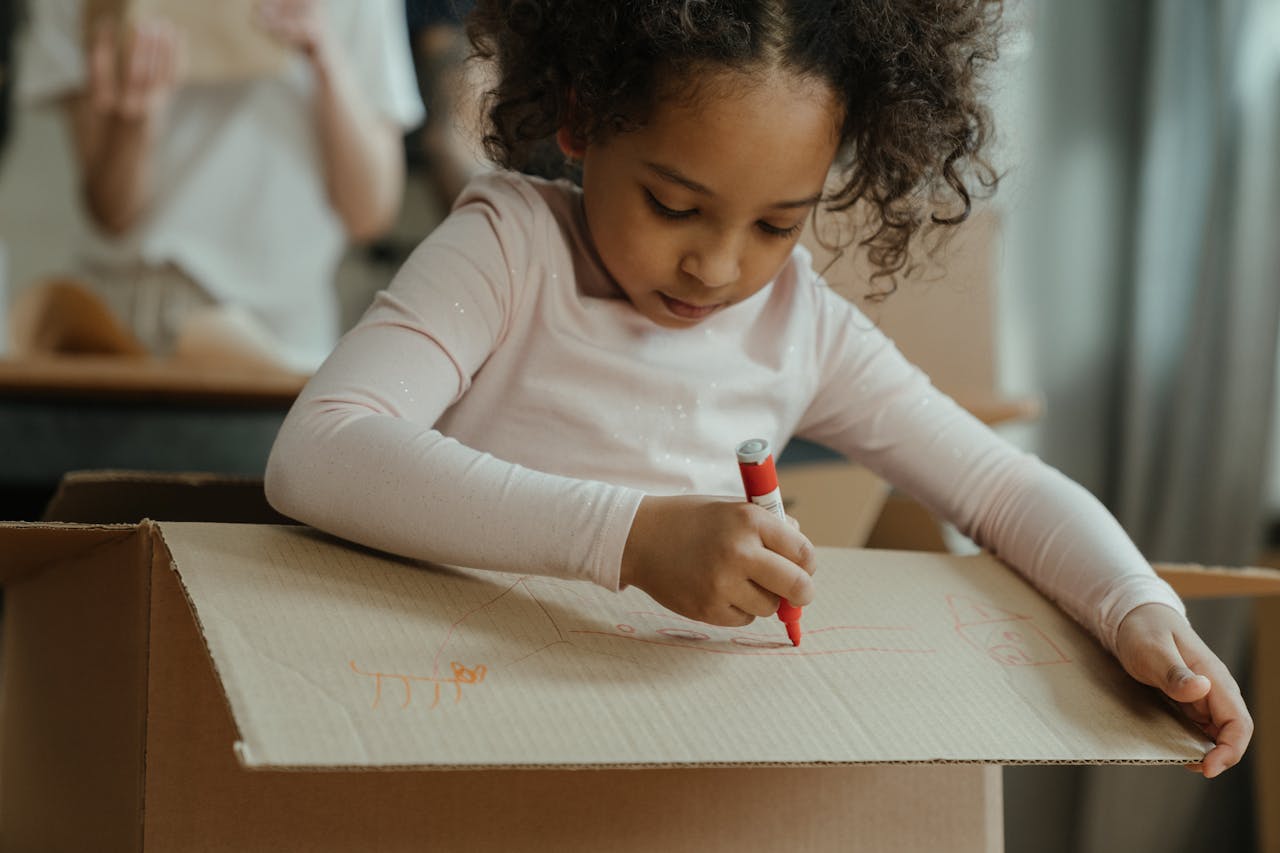 Young girl engrossed in drawing on a cardboard box during a creative indoor activity.